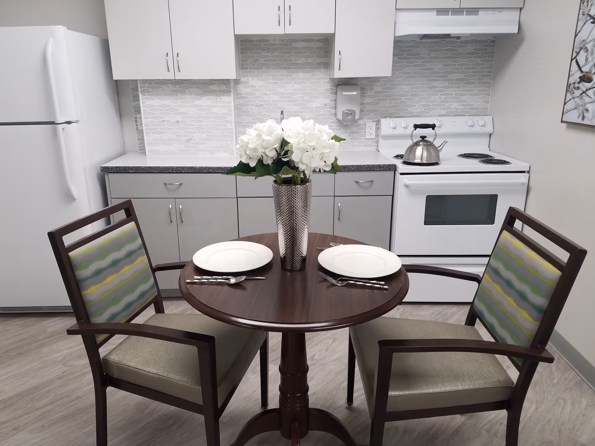 A small kitchen area with a round wooden table set for two with white plates, forks, and knives. A silver vase with white flowers is centered on the table. The kitchen includes a white refrigerator, white cabinets, a white stove with a kettle on it, and a gray countertop with a tiled backsplash. Two wooden chairs with patterned cushions are placed at the table.