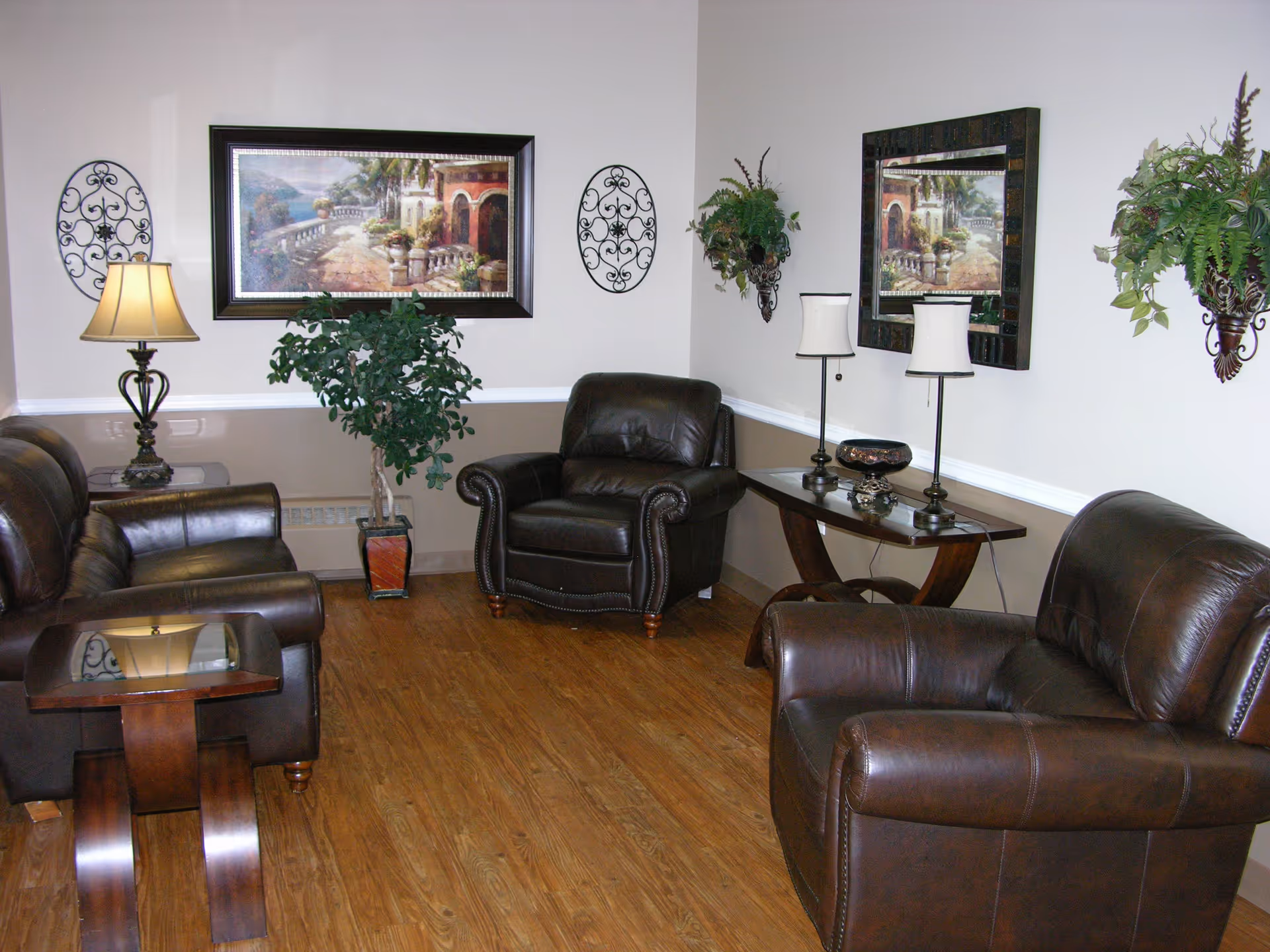 A cozy living room with three dark brown leather armchairs and a matching sofa arranged around wooden side tables. The room features wood flooring, beige walls with white trim, decorative wall art including a large framed painting and two metal wall hangings, and two wall-mounted planters with green foliage. There are also two table lamps on a wooden console table against the wall.