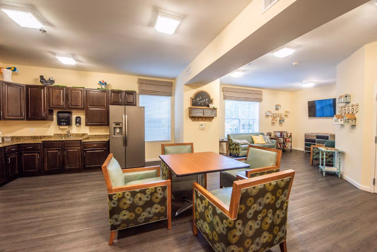 A bright senior living common room with a kitchenette, a central dining table surrounded by patterned chairs, and a seating area with sofas and a wall-mounted TV.