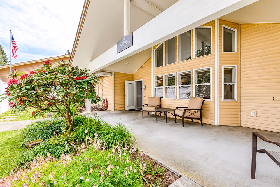 Outdoor covered patio area at The Cottages of Lynnwood with two cushioned chairs and a small table against a beige building wall with multiple windows. There is a garden with green plants and pink flowers in the foreground, and an American flag on a flagpole in the background.