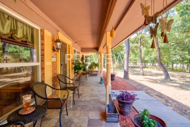 A covered outdoor porch area with several black metal chairs and small tables. There are potted plants and decorative hanging butterflies. The porch overlooks a yard with trees and a walking path.