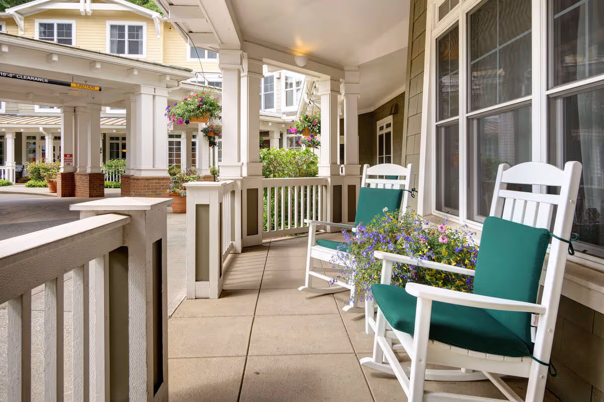Covered porch area with two white rocking chairs featuring green cushions, a flower pot with purple and yellow flowers between the chairs, hanging flower baskets, and a view of the building exterior with windows and columns.