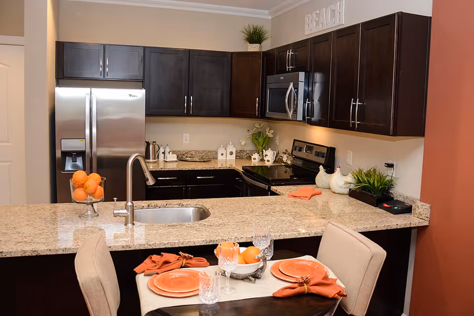 Modern kitchen with dark wood cabinets, stainless steel refrigerator, microwave, and stove. Granite countertops with a sink and a small dining table set with orange plates, napkins, and glassware. Decorative plants and ceramic chickens are placed on the counter, with a sign that reads 'BEACH' on top of the cabinets.
