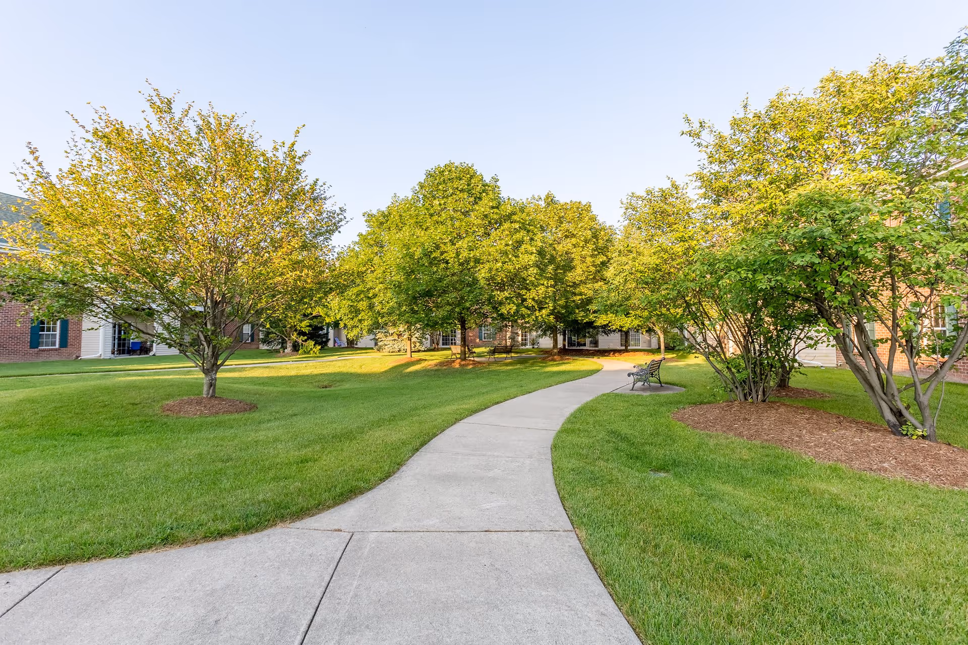 A curved concrete pathway winds through a well-maintained grassy area with several trees and shrubs. There is a bench along the path, and residential buildings with brick and siding exteriors are visible in the background under a clear blue sky.
