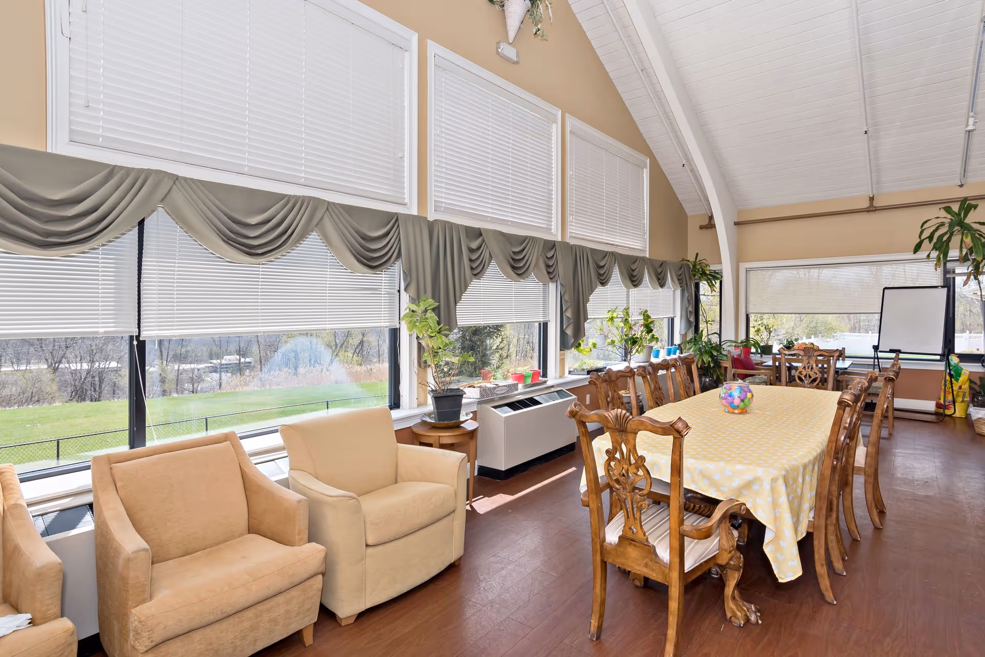 A bright dining room with large windows covered by white blinds and gray valances. The room features a long wooden dining table covered with a yellow tablecloth and surrounded by wooden chairs. There are two beige armchairs near the windows and several potted plants placed on tables and window sills. The ceiling is vaulted and painted white, and there is a whiteboard on a stand in the corner.