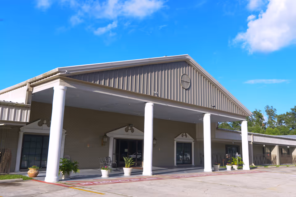 Exterior front view of Pontchartrain Health Care Center building with a covered entrance supported by white columns, potted plants near the entrance, and a clear blue sky with some clouds above.