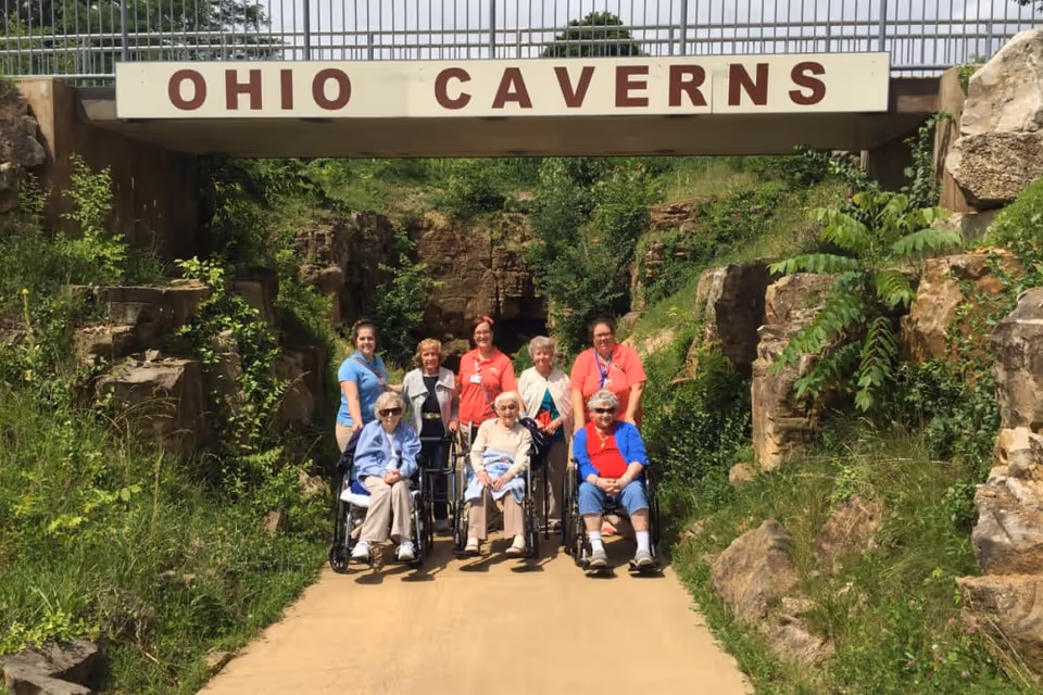 A group of elderly women, some in wheelchairs, accompanied by caregivers, posing for a photo in front of a rock formation entrance labeled 'OHIO CAVERNS' surrounded by greenery.