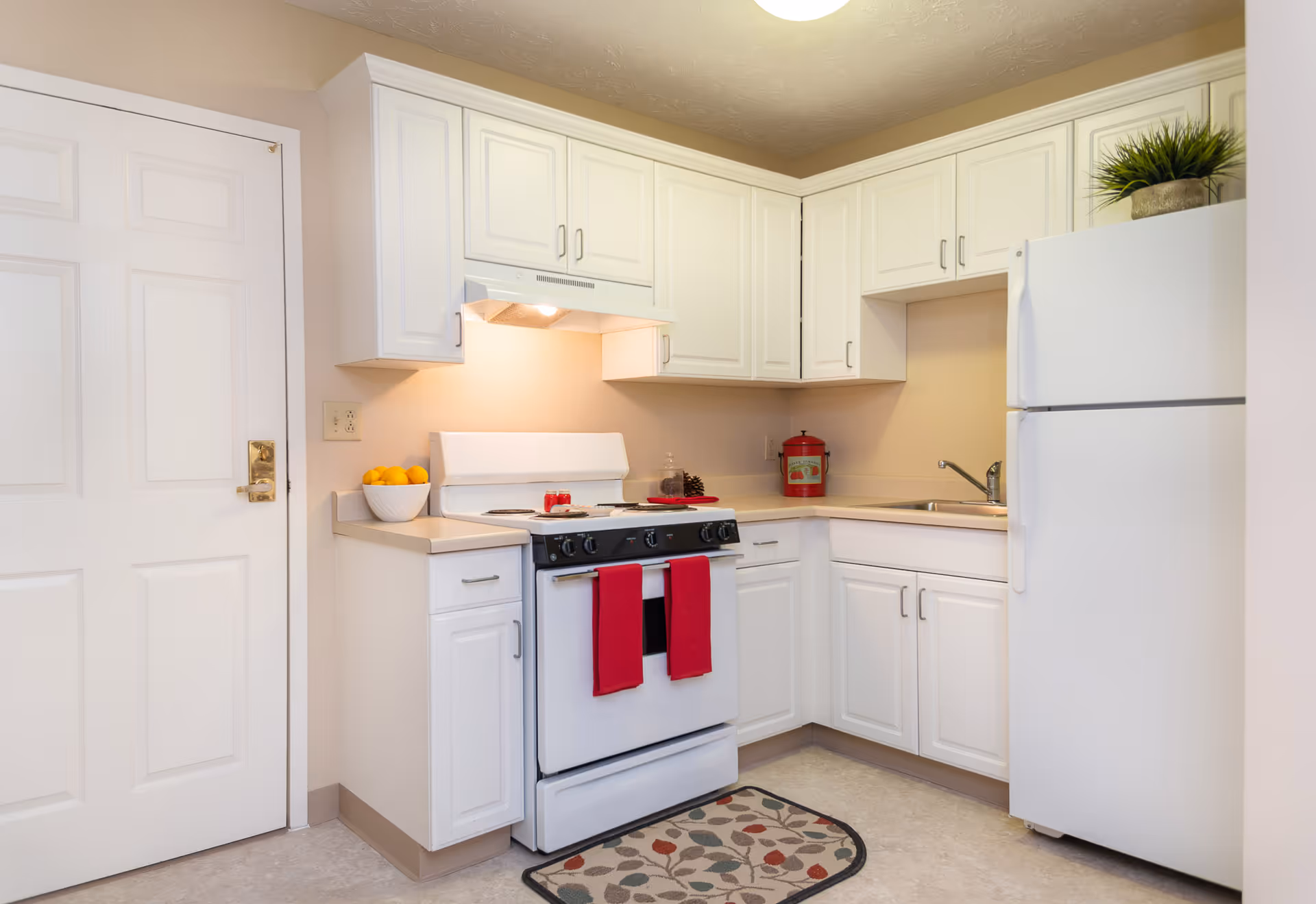 Bright compact kitchen with white cabinets, a stove with red towels, a refrigerator, sink, and countertop decor.