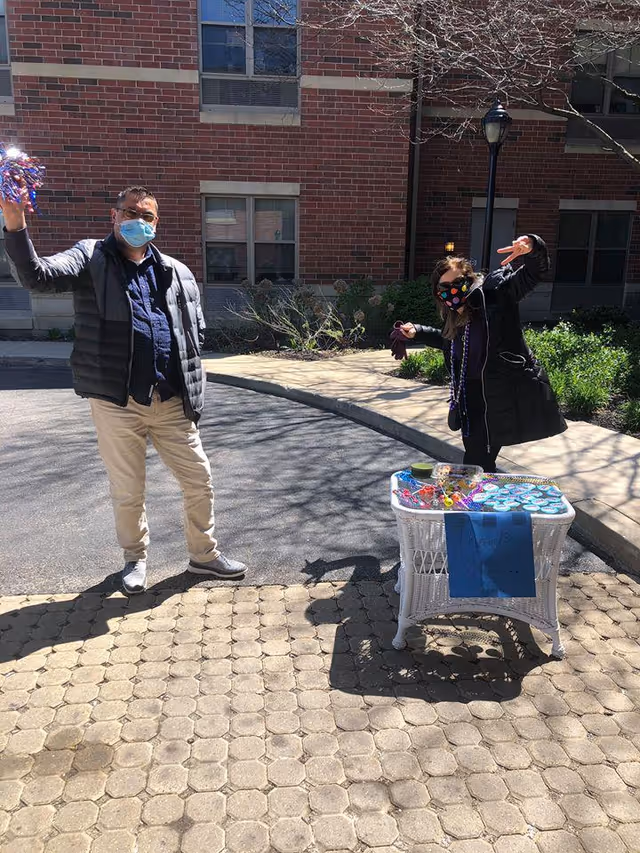 Two people wearing face masks stand outside near a brick building. One person on the left is holding up pom-poms, and the other person on the right is making a peace sign gesture. A small white wicker table with various items and a blue sign is in front of them on a paved area.