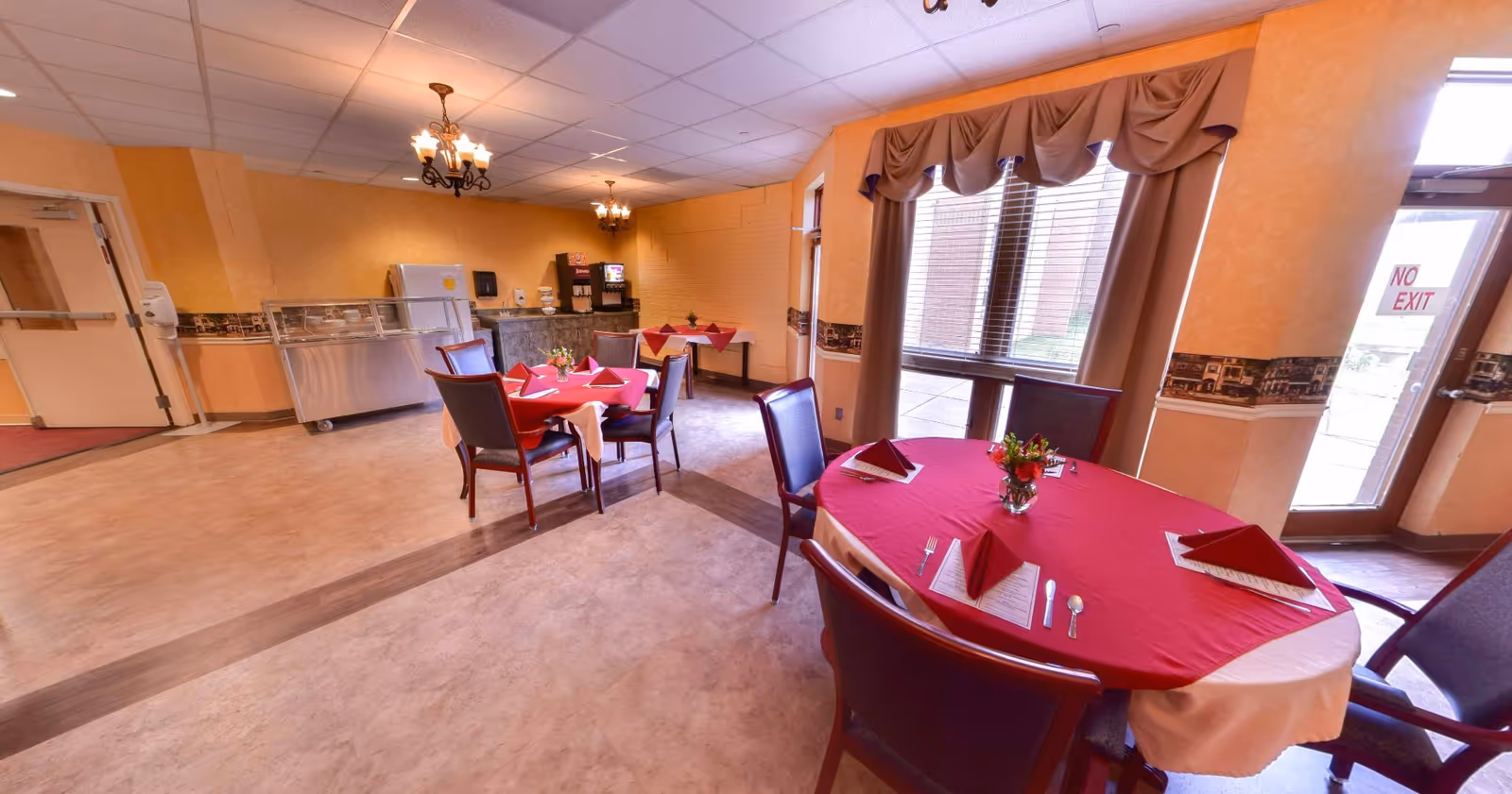 Dining room with round tables covered in red tablecloths and place settings, a buffet counter and beverage station in the background.