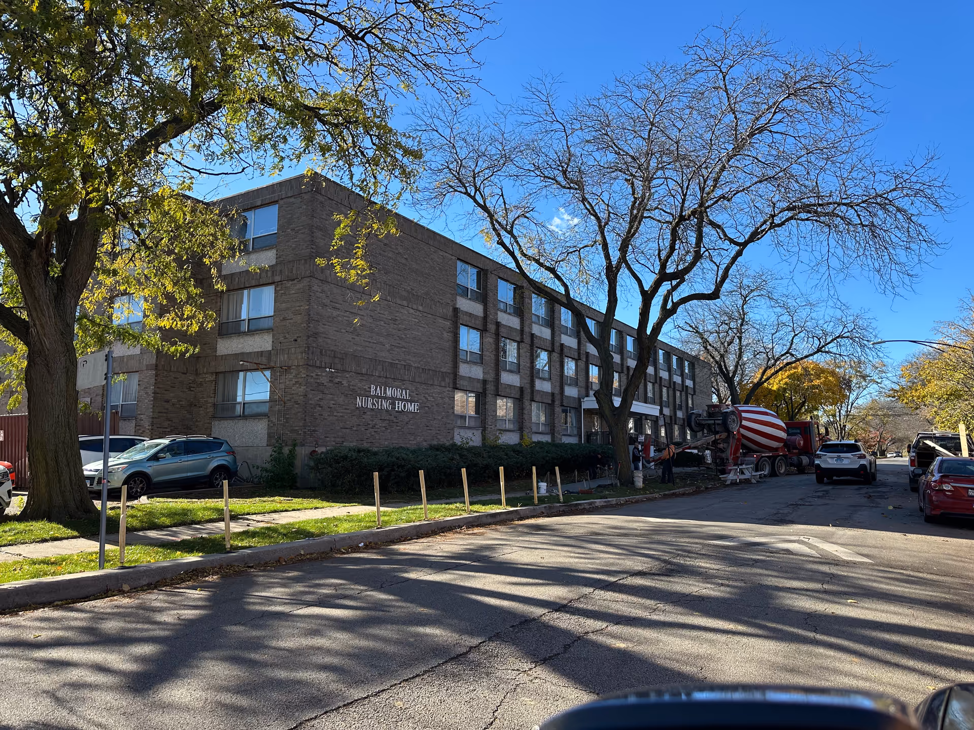 Exterior view of Balmoral Nursing Home, a three-story brick building with multiple windows. There are trees with some autumn leaves and several parked cars along the street. A cement mixer truck and workers are visible near the building entrance.
