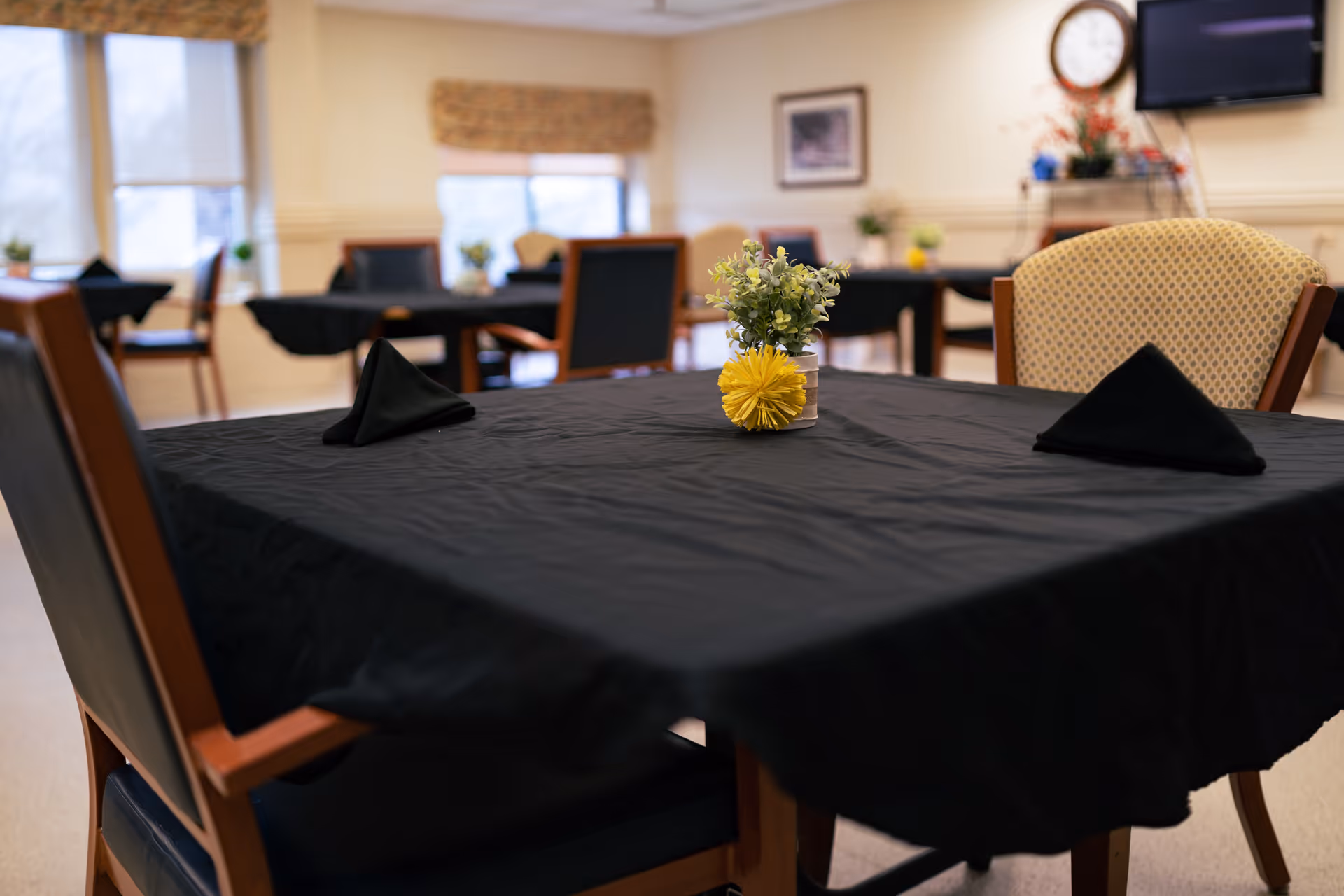 A dining room in a senior living facility with tables covered in black tablecloths, each set with black folded napkins and small floral centerpieces. Chairs with wooden frames and cushioned seats surround the tables. The room has large windows with beige valances, a wall clock, and a mounted television.