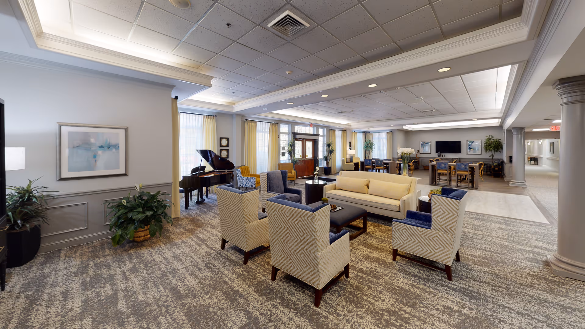 A spacious and well-lit living room area in a senior living facility featuring a beige sofa, four patterned armchairs with blue cushions arranged around a dark coffee table, a grand piano near the windows with yellow curtains, several potted plants, framed artwork on the walls, and a dining area with tables and chairs in the background.
