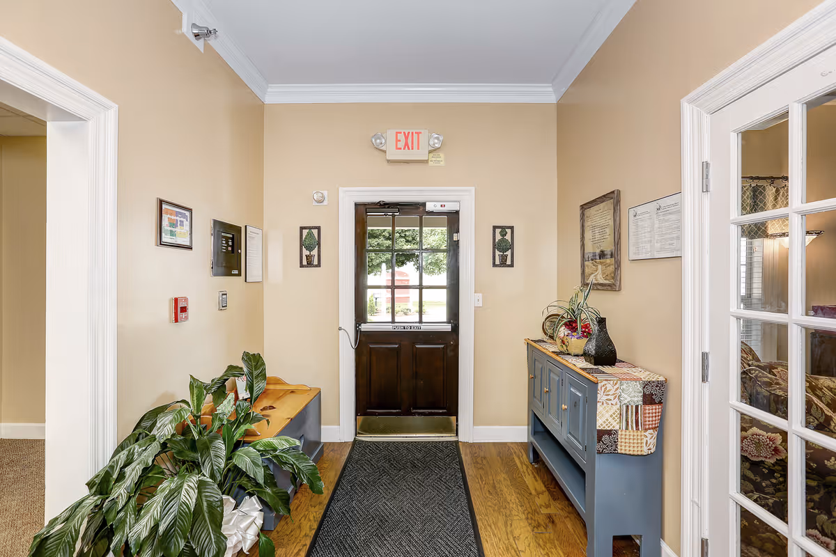 Interior view of an entryway in a senior living facility with a wooden door featuring glass panels and an exit sign above it. The walls are painted beige, and there is a blue console table with decorative items and a plant on the right side. On the left side, there is a wooden bench with a large green plant next to it. The floor is wooden with a dark rug leading to the door. A room with a glass-paneled door is visible on the right.