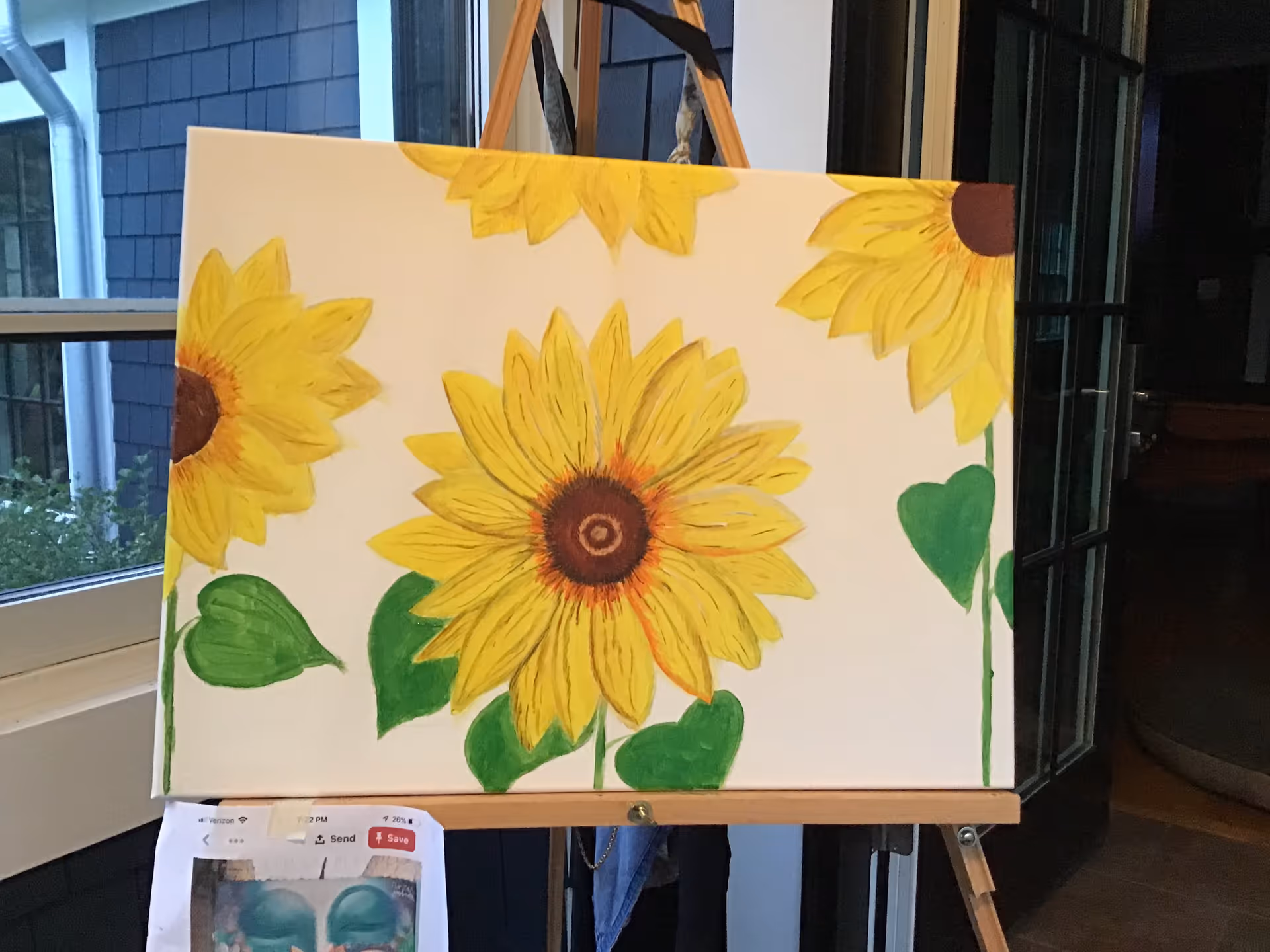 A sunflower painting on an easel displayed indoors next to a window.