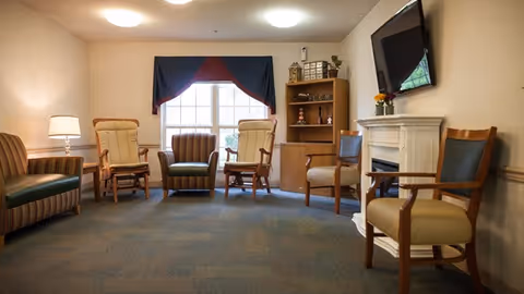 A cozy living room area with several wooden armchairs arranged around a window with a dark valance. There is a striped sofa on the left, a wooden cabinet with decorative items, a white fireplace with a flat-screen TV mounted above it, and a carpeted floor.