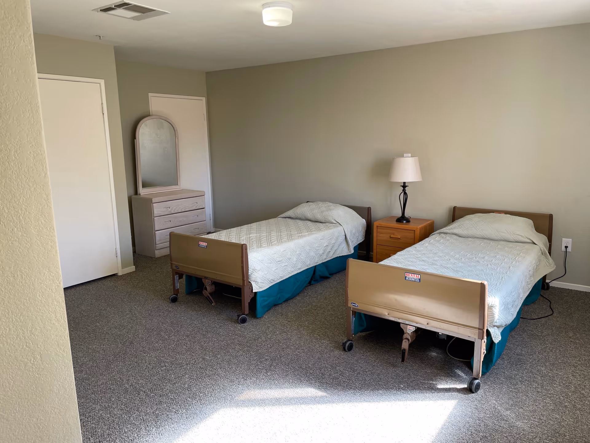 A simple bedroom with two single hospital-style beds with white bedspreads and teal bed skirts. Between the beds is a wooden nightstand with a lamp. In the corner, there is a dresser with a mirror. The room has beige walls and carpeted floor with sunlight coming through a window.