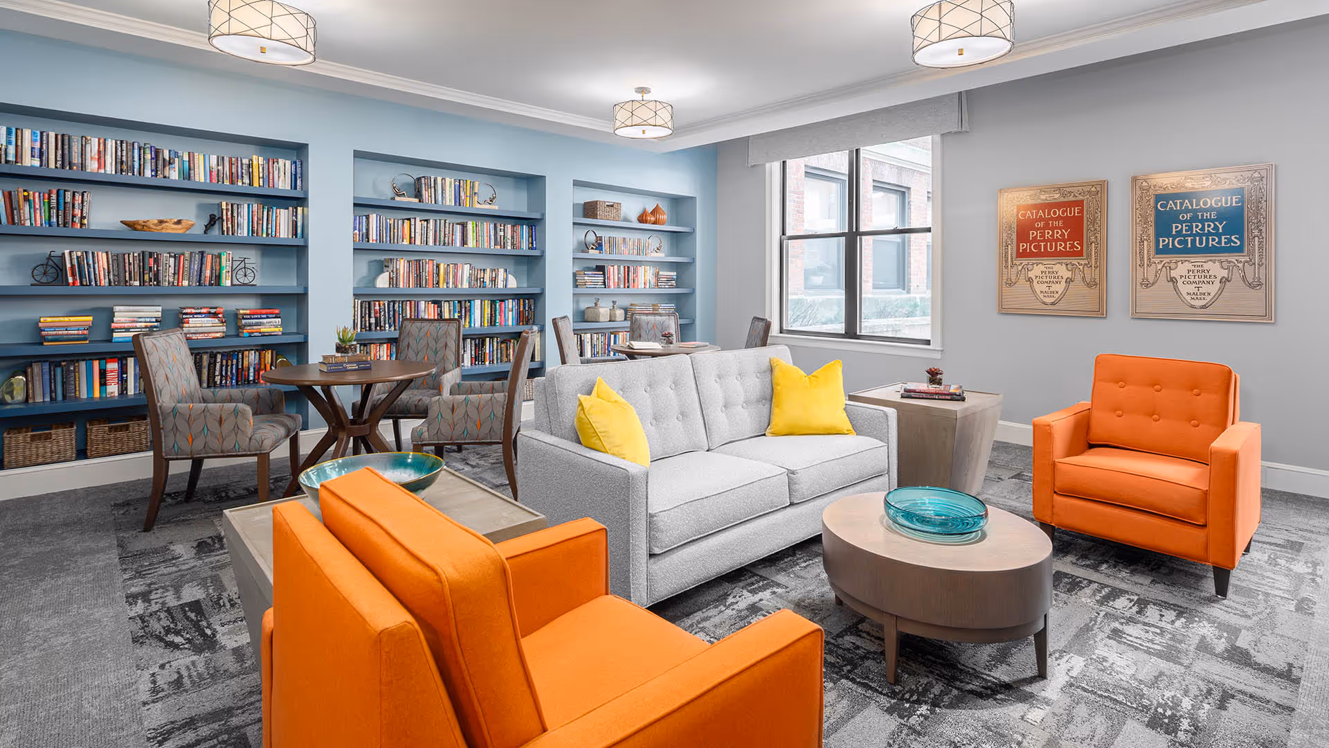 A bright and modern common area with a gray sofa adorned with yellow pillows, two orange armchairs, a round coffee table with a decorative blue bowl, and a small round table with four patterned chairs. The back wall features built-in blue bookshelves filled with books and decorative items. Two framed vintage-style posters hang on the adjacent wall near a large window letting in natural light.