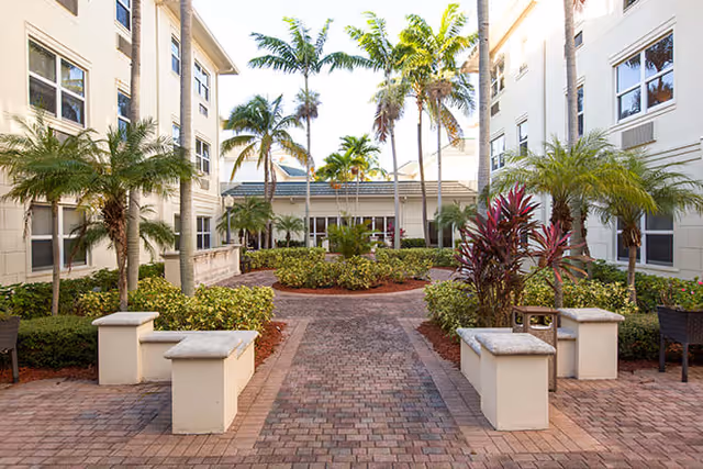 Outdoor courtyard area at Brookdale Jensen Beach featuring a brick pathway lined with benches and lush tropical plants including palm trees and shrubs, surrounded by light-colored multi-story buildings.