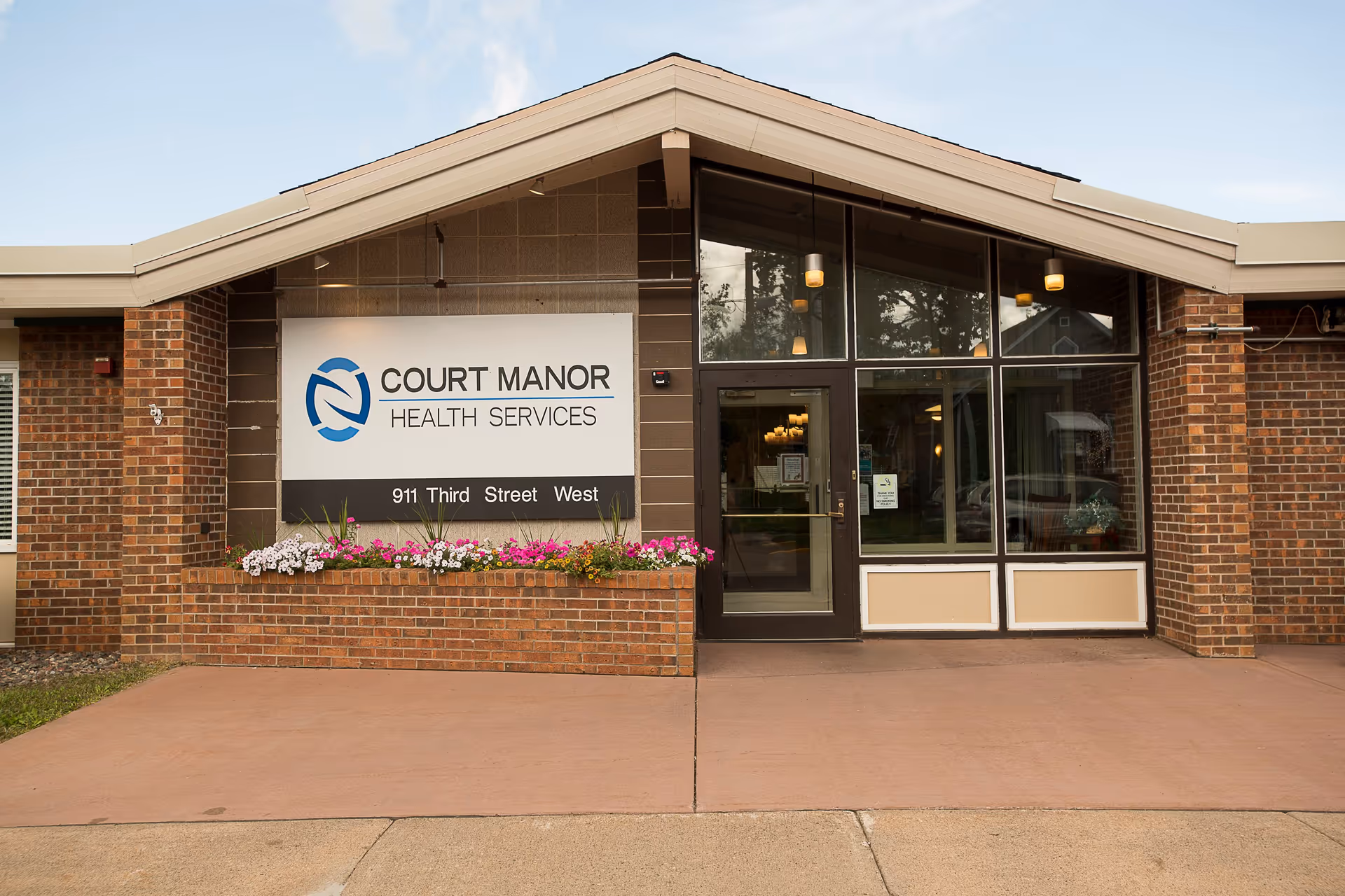 Front entrance of Court Manor Health Services building with a large sign displaying the facility name and address 911 Third Street West. The entrance features glass doors and windows, brick walls, and a flower bed with colorful flowers beneath the sign.