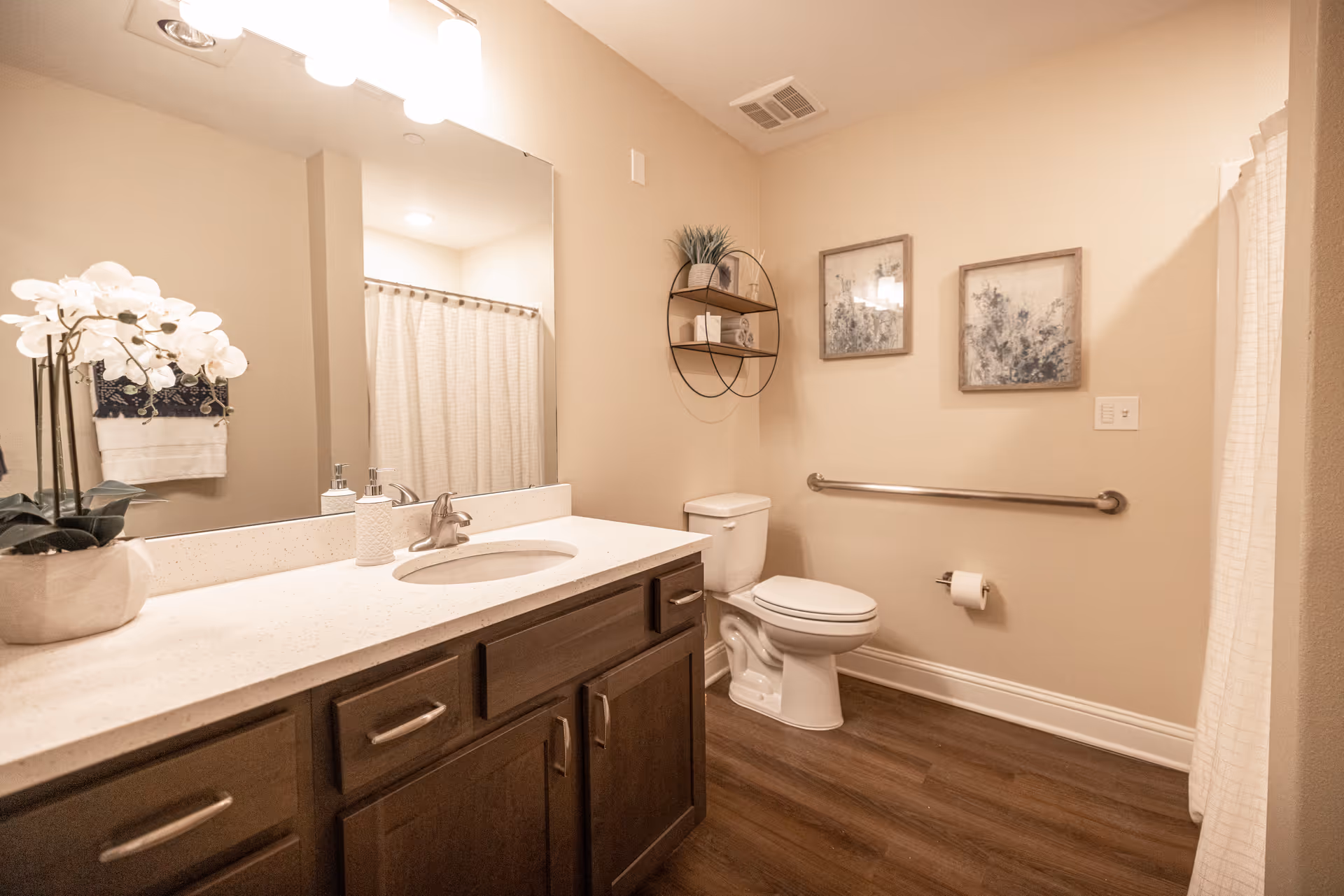 A clean and well-lit bathroom featuring a white countertop with a sink, a large mirror above the sink, a toilet with a grab bar on the wall beside it, a shower with a curtain, wooden cabinetry below the sink, decorative wall art, and a small round shelf with plants and decor items.