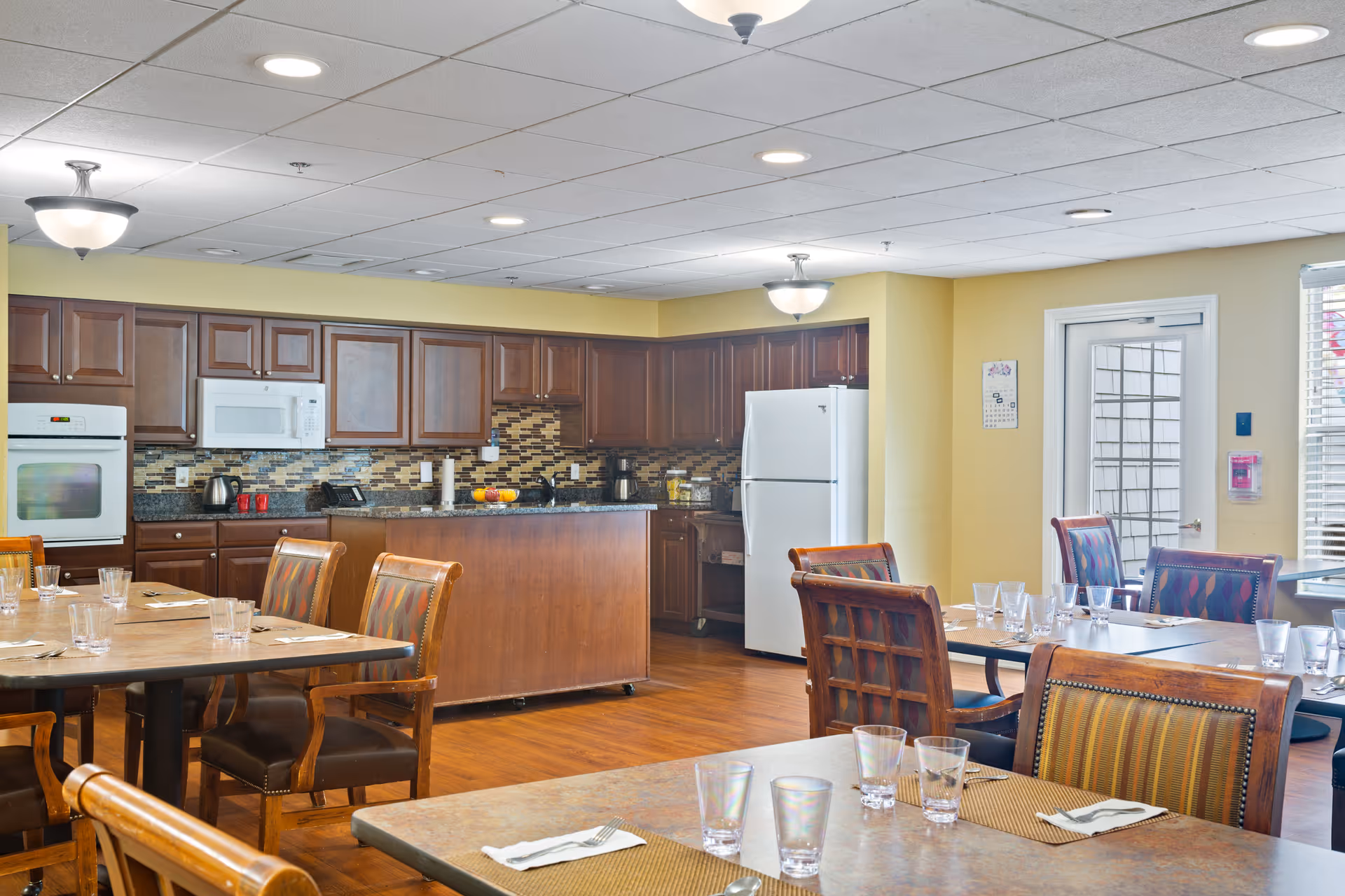 A bright dining area with multiple tables and chairs set with glasses and napkins. In the background, there is a kitchen with wooden cabinets, a white refrigerator, a microwave, an oven, and a kitchen island. The room has a yellow wall and wood flooring, with ceiling lights providing illumination.
