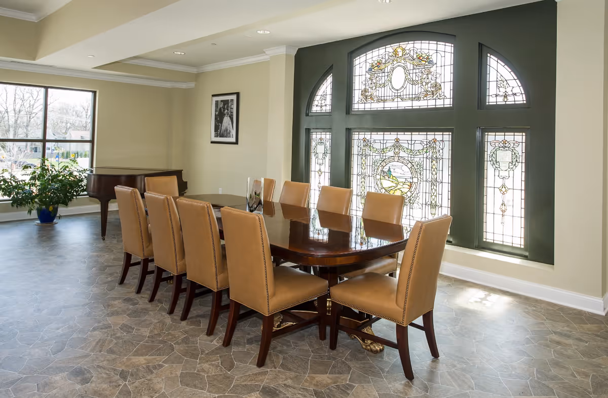 A dining room with a long wooden table surrounded by ten tan upholstered chairs. The room features a large stained glass window with intricate designs, a grand piano in the corner, a potted plant near a large window, and a framed black and white photograph on the wall.