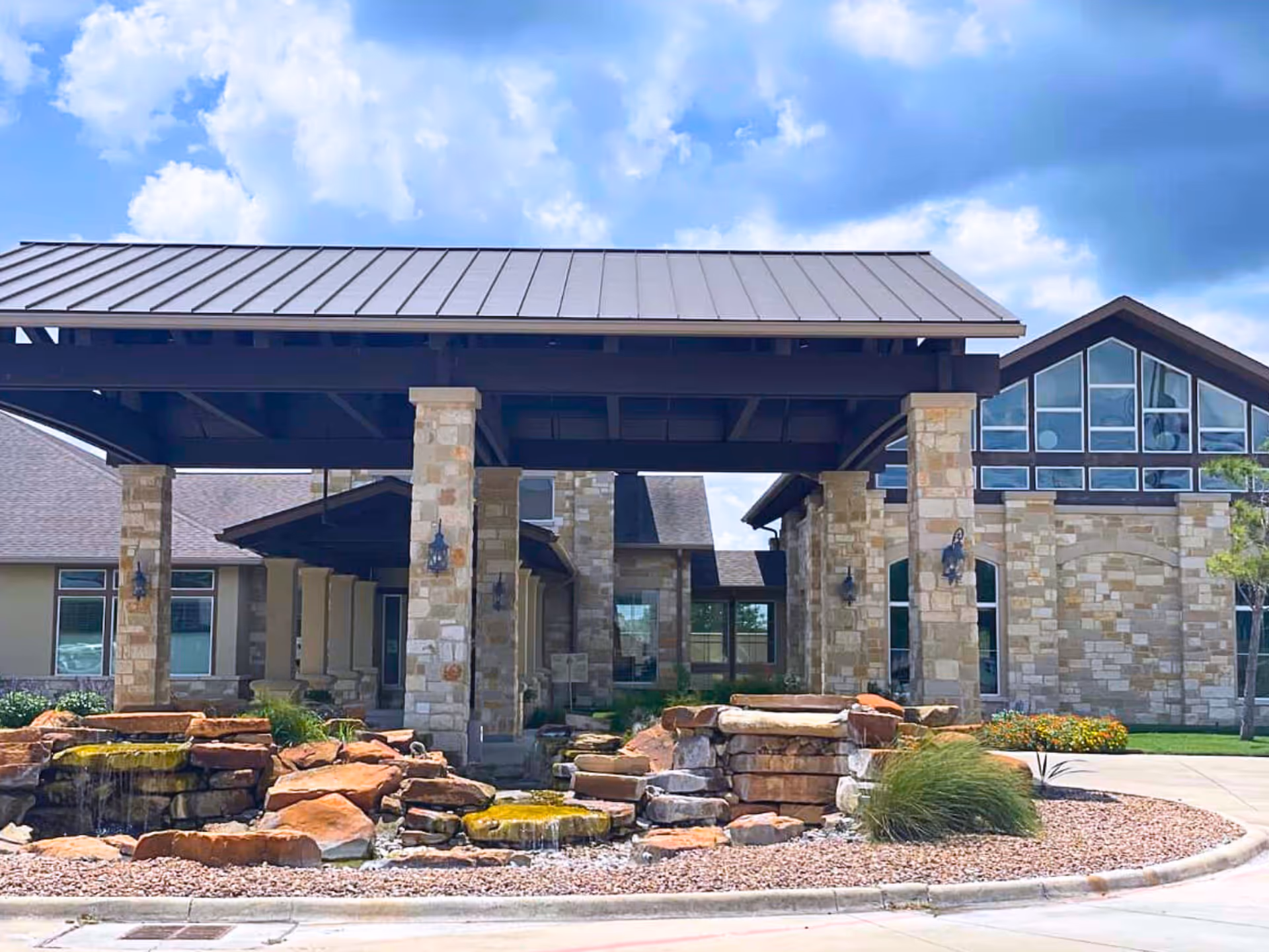 Front exterior view of North Twin Pines Nursing Home featuring a covered entrance with stone pillars, a landscaped rock water feature, and a building with large windows under a partly cloudy sky.