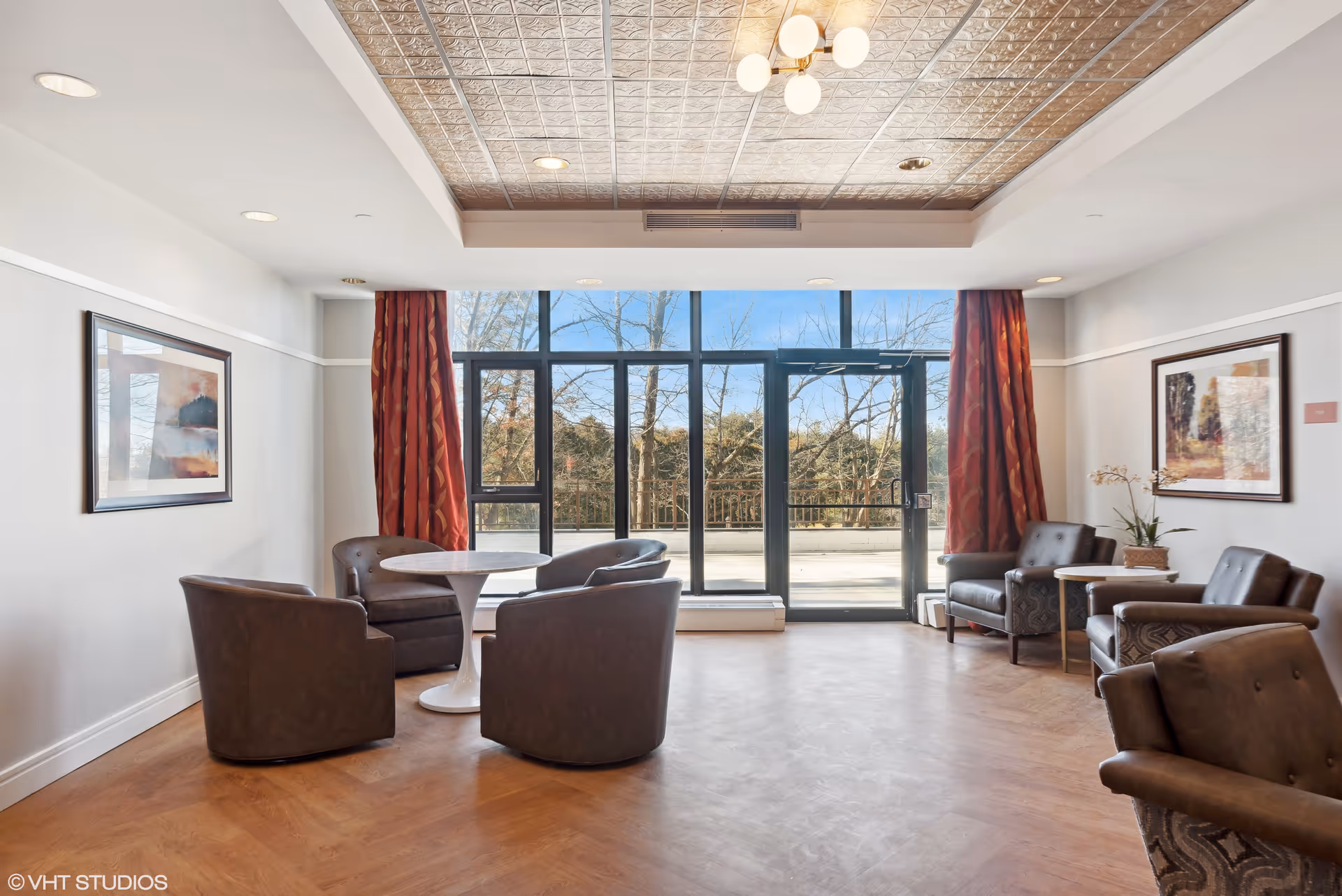 A bright and spacious lounge area with large floor-to-ceiling windows and a glass door leading outside. The room features a round white table surrounded by four brown leather chairs on the left, and two brown leather armchairs with a small side table and a potted plant on the right. The walls are decorated with framed artwork and red patterned curtains flank the windows. The ceiling has a decorative metallic tile design with recessed lighting and a modern chandelier.