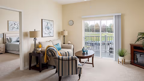 Sunlit living room featuring a sofa, striped armchair, coffee table, lamps, and sliding glass door to a balcony with a grassy view, with a bedroom visible through a doorway.