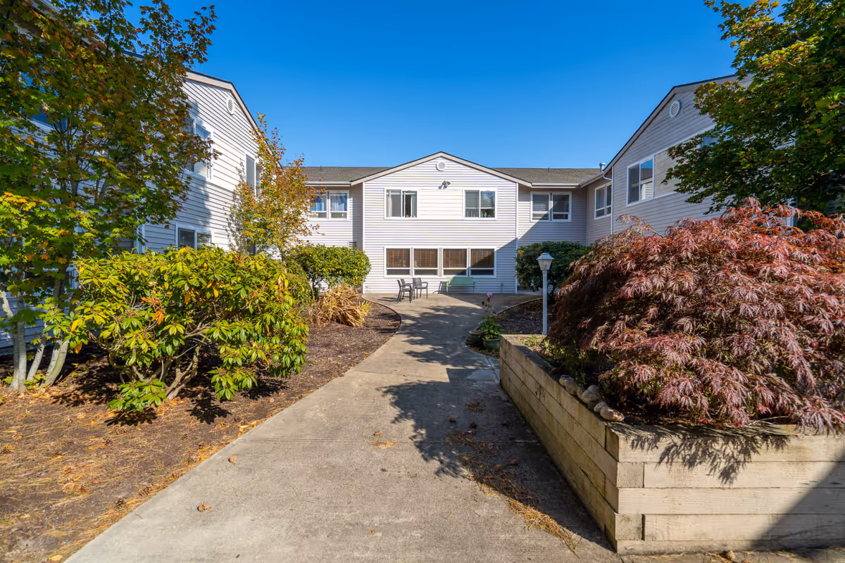 Courtyard walkway and landscaped entrance leading to the front of a two-story white retirement facility building under a clear blue sky.