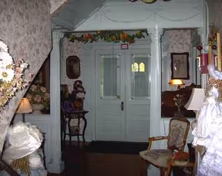 An ornate Victorian mansion foyer with white woodwork, garlands, lamps, and chairs around a central doorway.