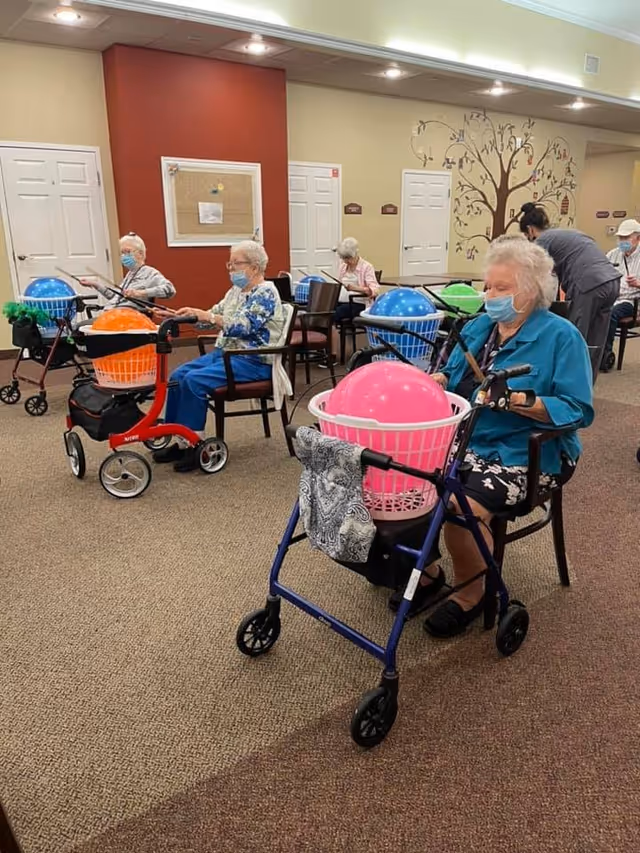 Several elderly women wearing masks are seated in a room, each with a walker in front of them holding a large colorful exercise ball in a basket. The room has beige walls, a red accent wall, and a decorative tree mural. Some other elderly individuals and a caregiver are visible in the background.