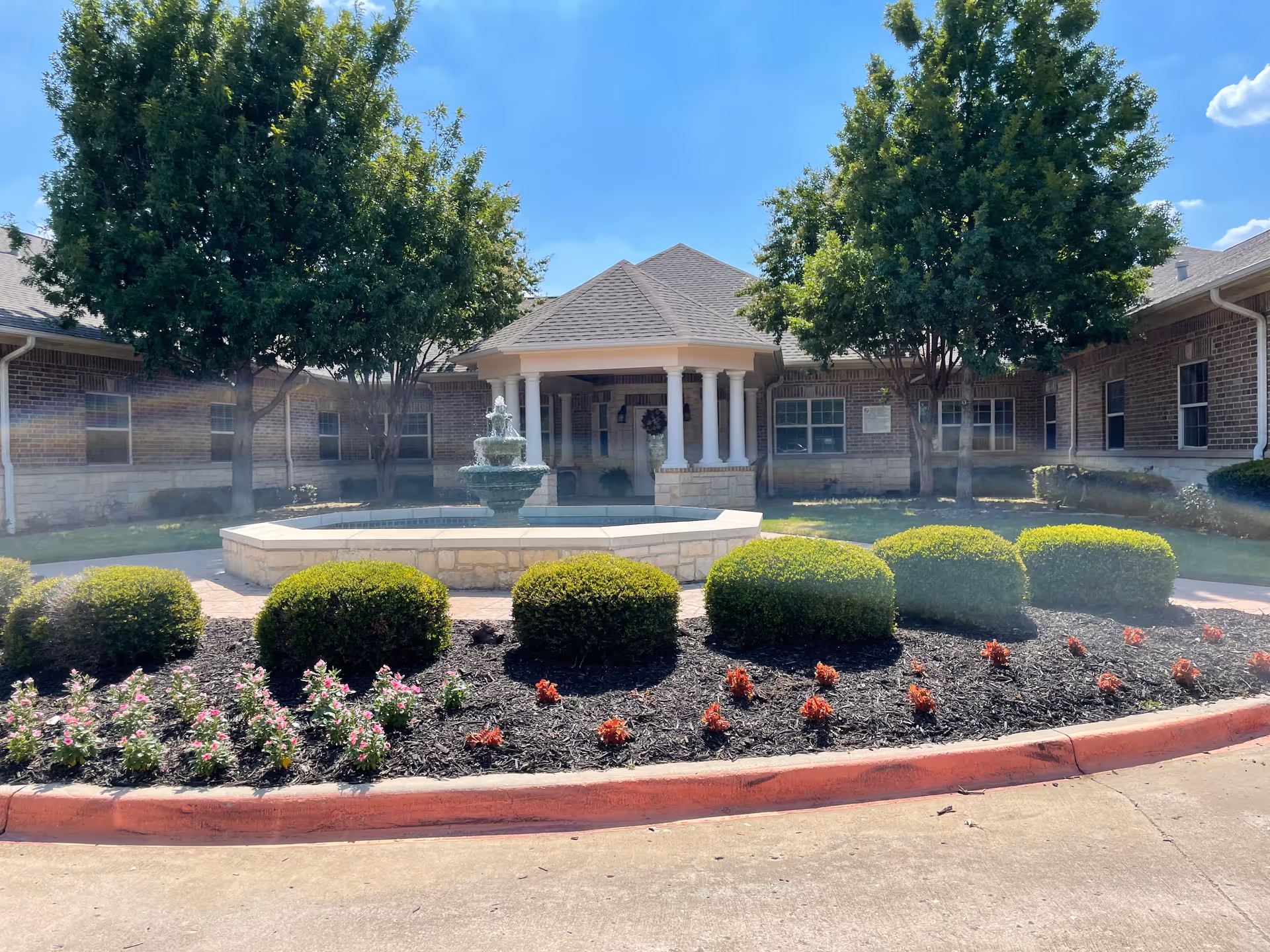 Front courtyard of Magnolia Assisted Living and Memory Care featuring a stone fountain surrounded by neatly trimmed bushes and flower beds, with a covered entrance supported by white columns and brick building walls under a clear blue sky.