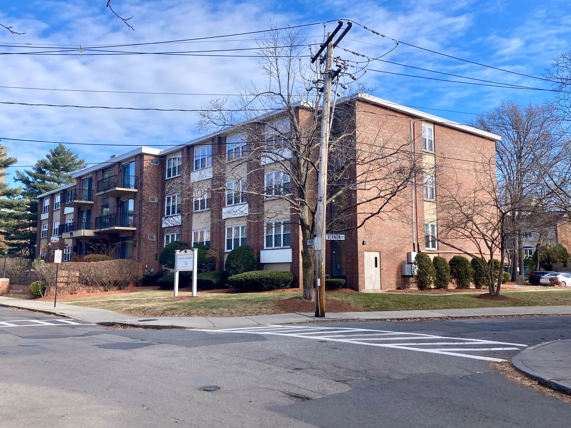 Exterior view of a three-story brick residential building with multiple windows and balconies, situated on a corner lot with a street sign showing Park and Vernon streets. The building is surrounded by trimmed bushes and trees without leaves, under a partly cloudy sky.