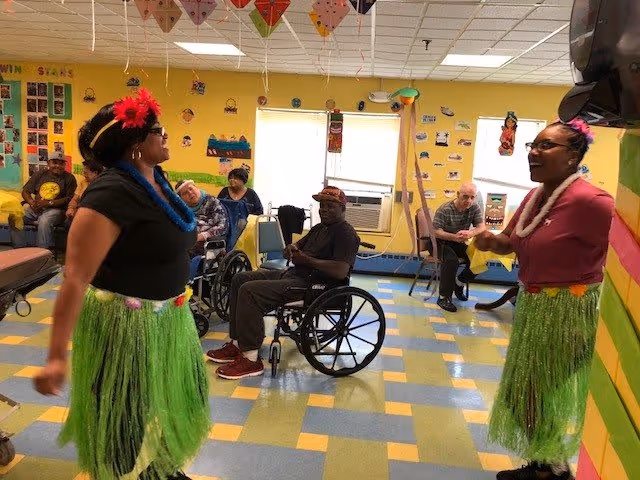 Two women dressed in Hawaiian-themed outfits with grass skirts and flower leis are dancing in a colorful room with yellow walls and a checkered floor. Several elderly people, some in wheelchairs, are seated and watching the performance. The room has decorations hanging from the ceiling and photos on the walls.
