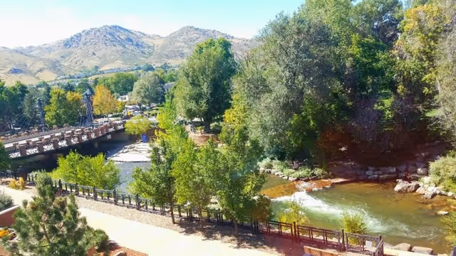 A scenic outdoor view featuring a flowing river bordered by trees and greenery, a pedestrian bridge crossing the river, and mountains in the background under a clear blue sky.