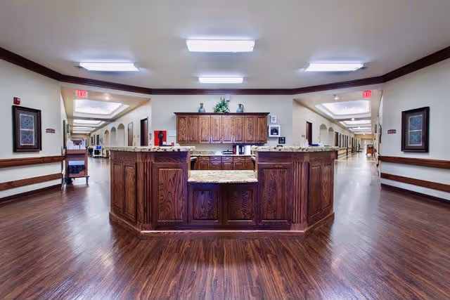 A spacious hallway in a senior care facility with a central wooden reception desk featuring granite countertops. The hallway has polished wooden floors, cream-colored walls with dark wood trim, framed artwork on the walls, and bright overhead lighting. Two long corridors extend from either side of the reception area.