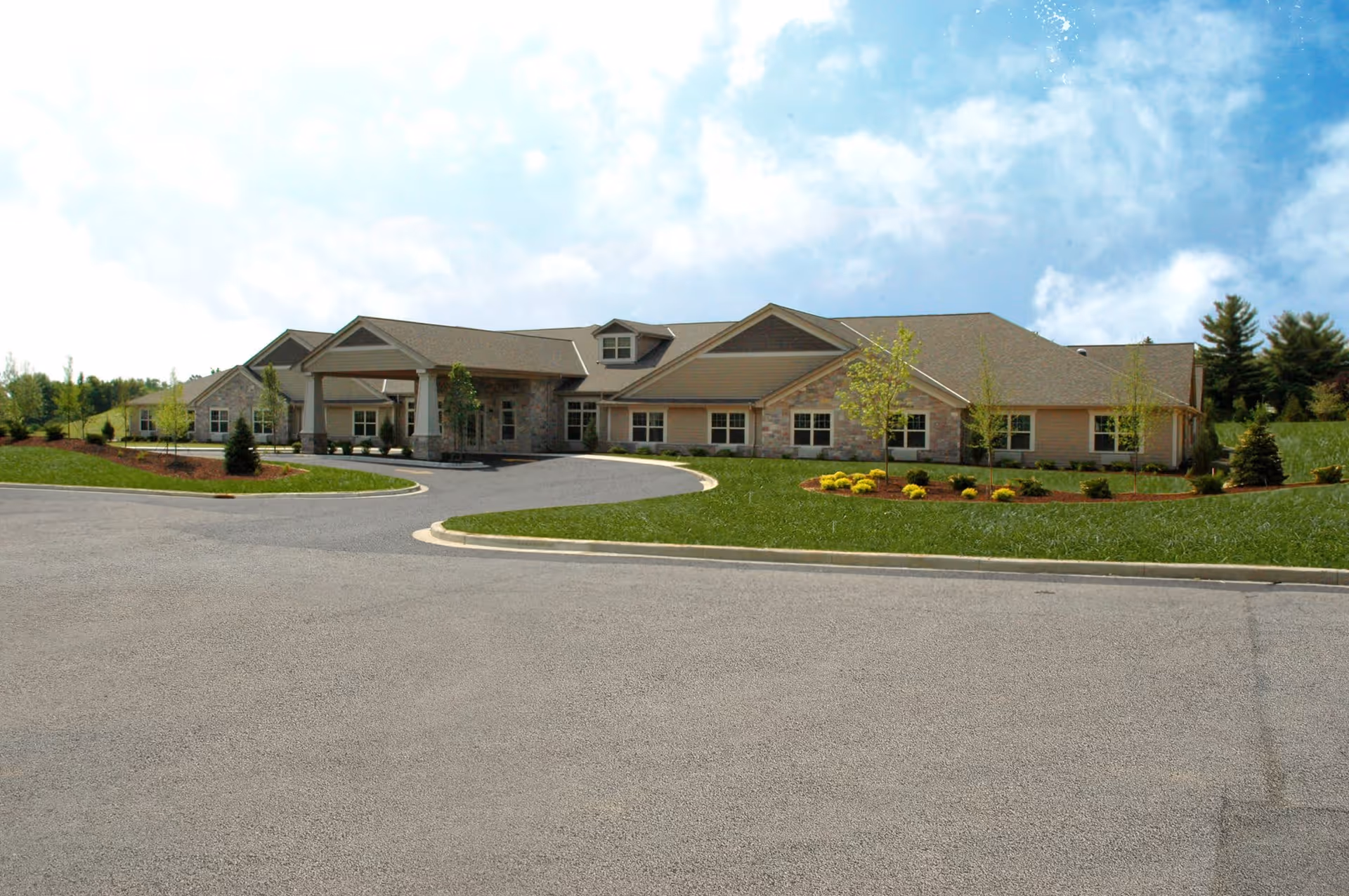 Exterior view of a single-story senior living facility building with a covered entrance, surrounded by landscaped greenery and a paved driveway under a partly cloudy sky.