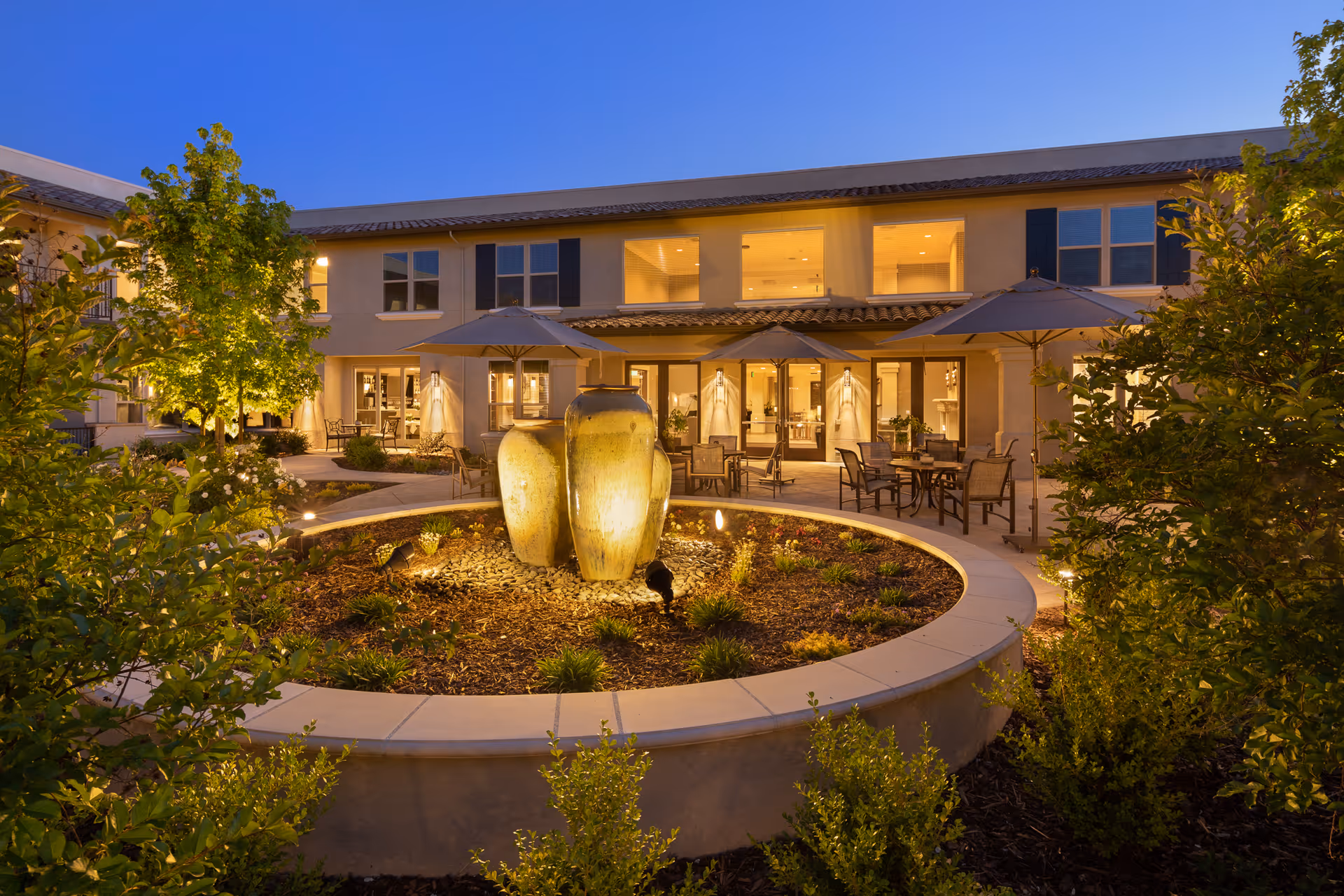 Evening view of an outdoor patio area at Oakmont of Westpark featuring a circular garden with three large decorative urns in the center, surrounded by greenery and shrubs. Several tables with chairs and umbrellas are arranged on the patio in front of a two-story building with illuminated windows and doors.