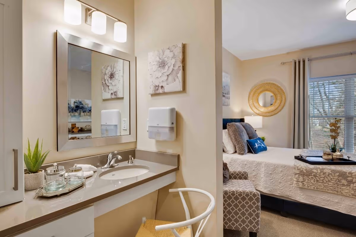 A cozy senior living space featuring a small vanity area with a sink, mirror, and light fixture on the left, and a bedroom area with a neatly made bed, decorative pillows, a round wall mirror, and a window with curtains on the right.