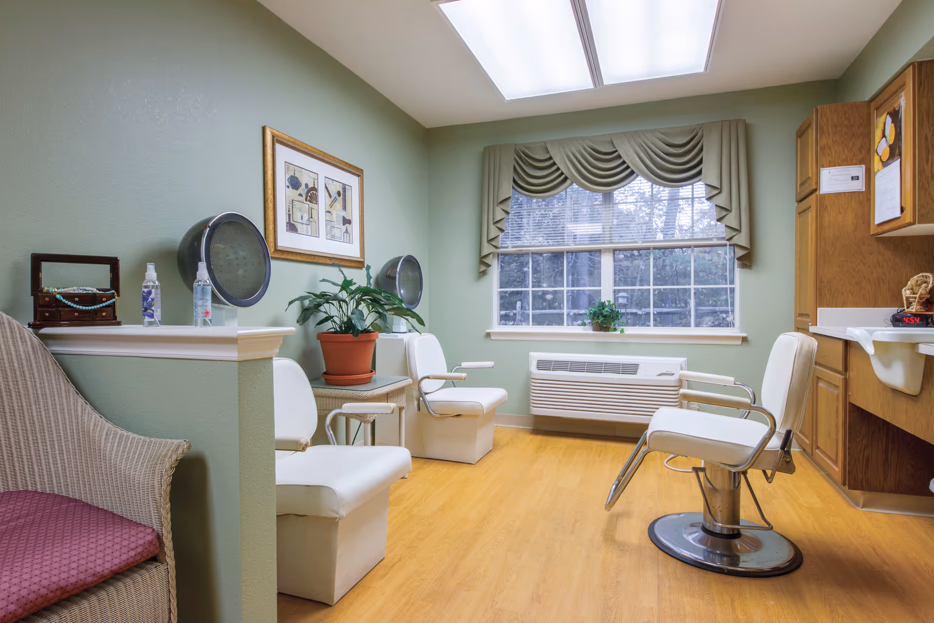 A small salon room with styling chairs, hooded dryers, a large window, potted plants and wooden cabinets.