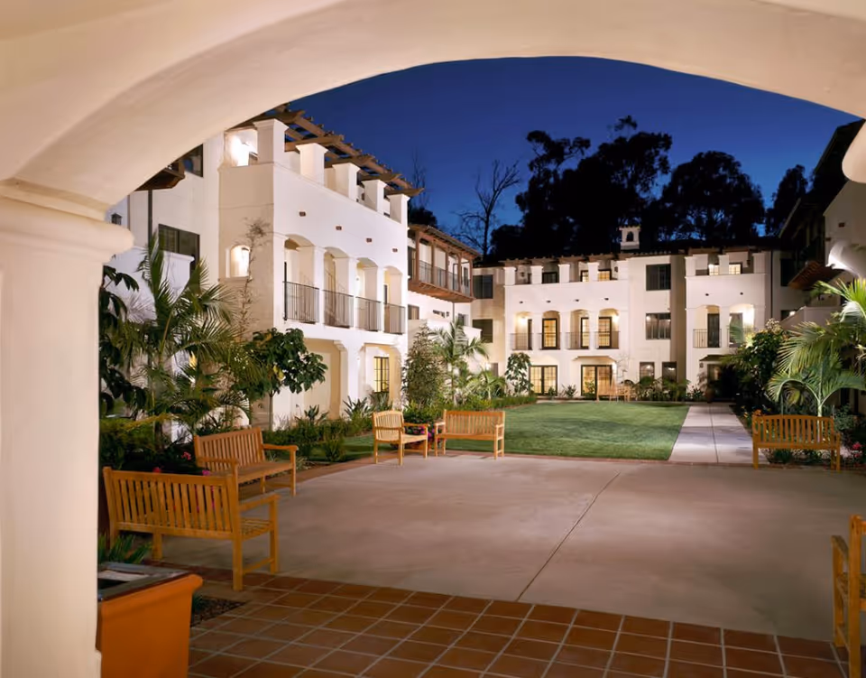View of a courtyard at night in a senior living facility named Maravilla, featuring white multi-story buildings with balconies, well-lit windows, green lawn, palm trees, and wooden benches arranged around the paved area.