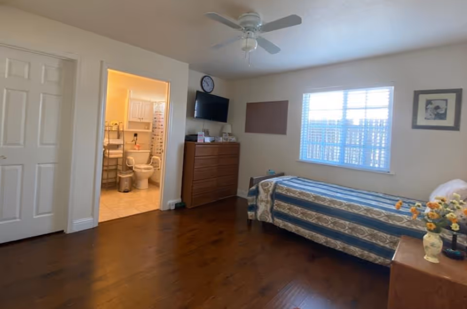 A bedroom with a single bed covered in a patterned blue and beige bedspread, a wooden dresser with a TV mounted above it, a ceiling fan, a window with blinds, and a door leading to a bathroom with a toilet and shower visible.