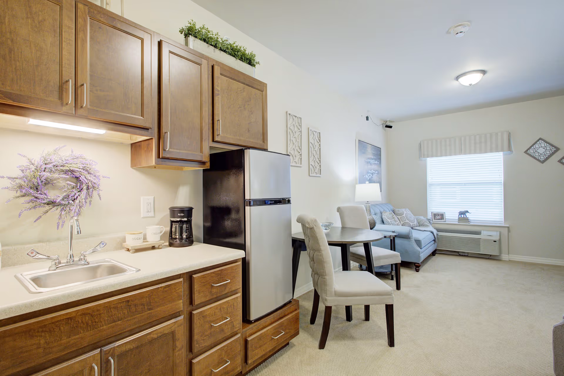 A bright and cozy living space featuring a small kitchen area with wooden cabinets, a stainless steel refrigerator, a coffee maker, and a sink. Adjacent to the kitchen is a round dining table with two upholstered chairs. In the background, there is a comfortable sofa with decorative pillows, a side table with a lamp, wall art, and a window with blinds and a valance. The room has light-colored walls and carpeted flooring.
