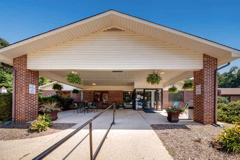 Covered entrance of a brick building with a white roof and hanging plants, featuring a ramp and seating area near the entrance doors under a clear blue sky.