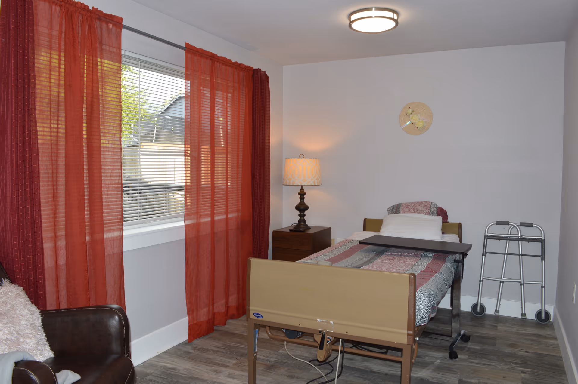 A small bedroom with a single hospital-style bed covered with a patterned quilt. There is a bedside table with a lamp, a walker against the wall, a brown armchair with a pillow, and a window with red curtains letting in natural light.