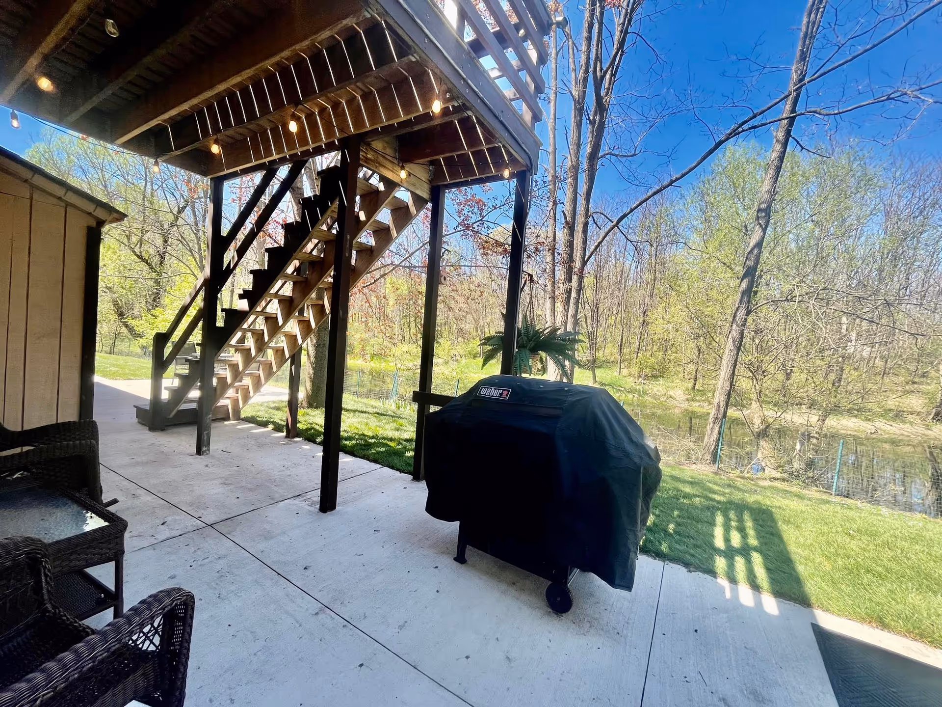Outdoor patio area with a covered grill, wicker chairs, a glass-top table, and a wooden staircase leading up to a balcony. The patio overlooks a grassy yard with trees and a small pond under a clear blue sky.