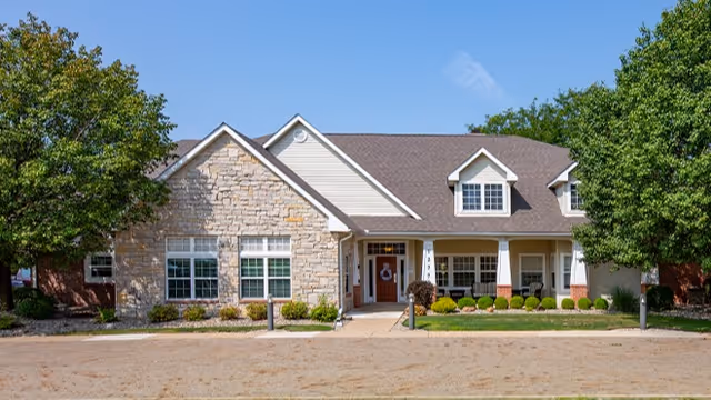 One-story senior living building with a stone and siding facade, central entrance and porch flanked by windows and trees.