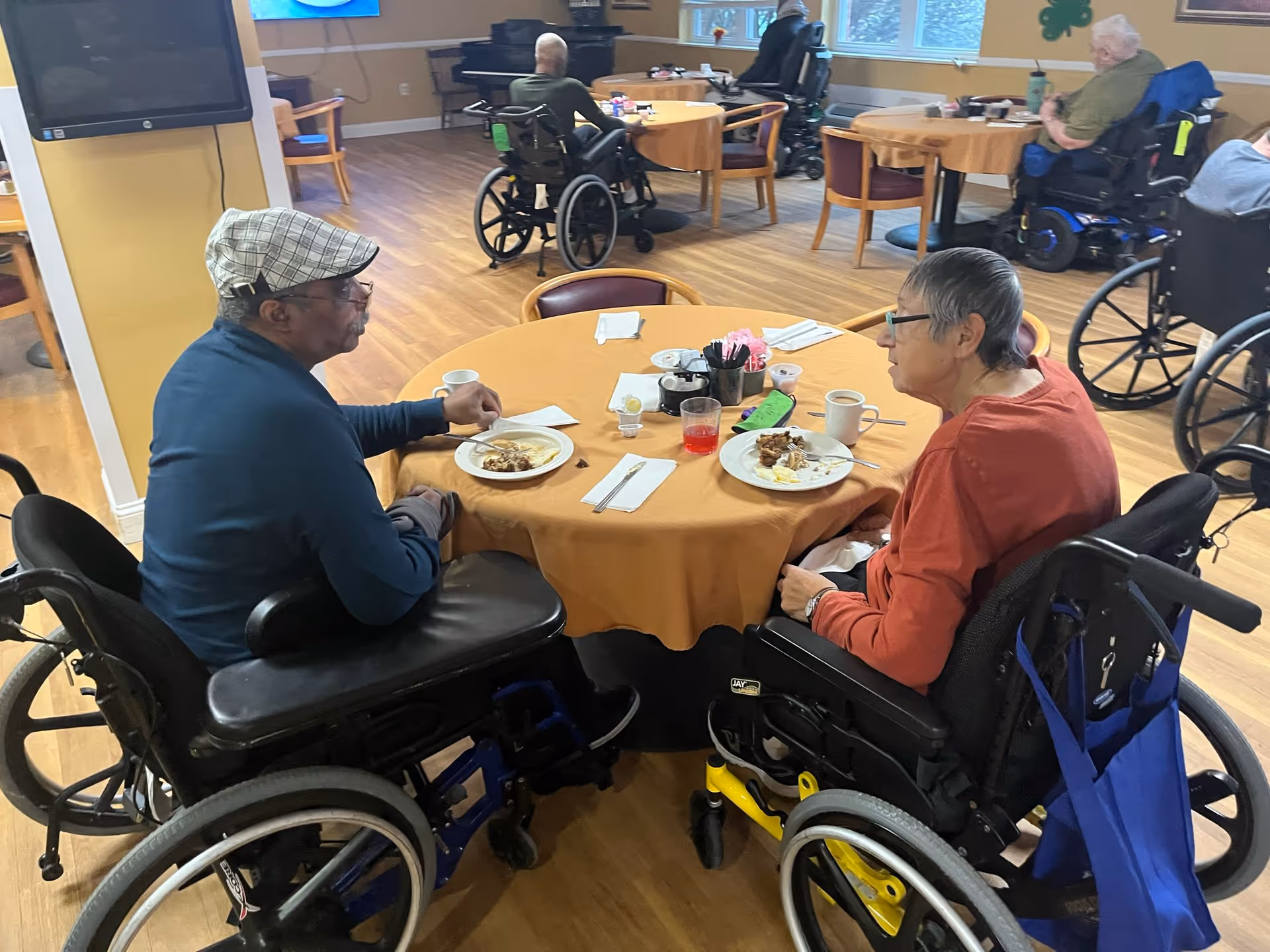 Two elderly individuals in wheelchairs sitting at a round table covered with a yellow tablecloth, eating a meal and conversing in a dining area with other people in wheelchairs seated at tables in the background.
